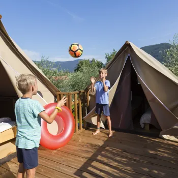 Two kids play with a ball on a wooden deck between two tents.