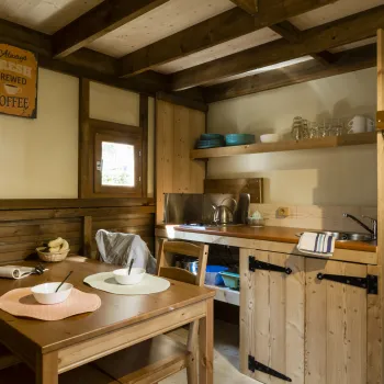 Cozy wooden kitchen with a table, fridge, and shelves of dishes. Warm lighting.