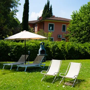 Lawn with lounge chairs and an umbrella near a red house surrounded by greenery.