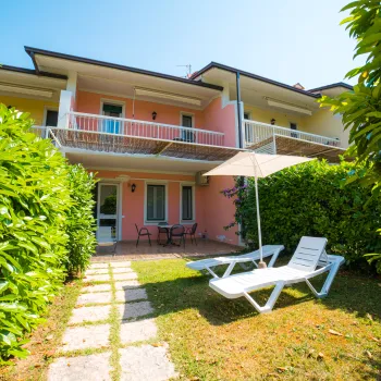 Pink house with balconies, surrounded by greenery, with two lounge chairs in the garden.