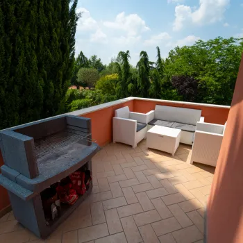 Sunny balcony with a grill, white seating, and lush greenery in the background.