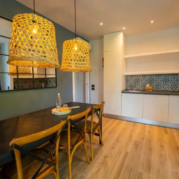 Dining room with wooden table, wicker chairs, and pendant lights; modern kitchen in background.