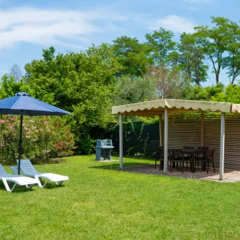 Sunny garden with a gazebo, lounge chairs, and a blue umbrella on grass.