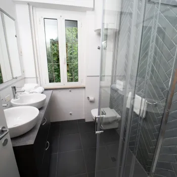 Modern bathroom with double sinks, glass shower, and window view of greenery.