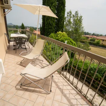 Sunny balcony with lounge chairs and umbrella, overlooking a garden and houses.