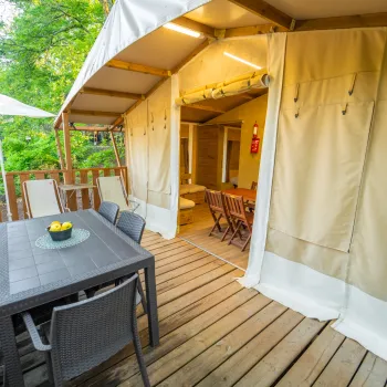 Wooden deck with a table, chairs, and beige tent; green foliage background.