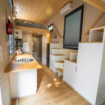 Modern tiny house interior with wooden floors, compact kitchen, stairs, and natural light.