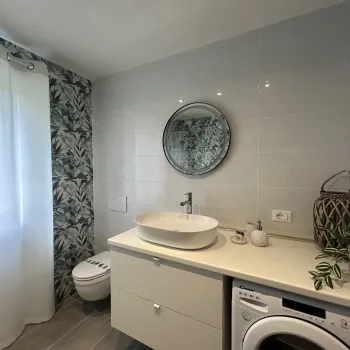 Bathroom with sink, round mirror, washing machine, floral wallpaper, and sheer curtains.