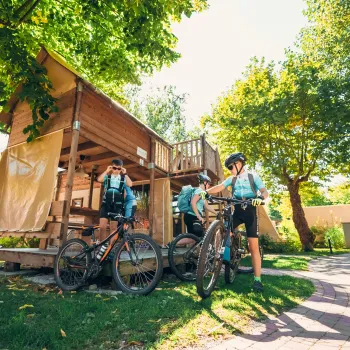 Cyclists with bikes near a wooden cabin in a sunny park.