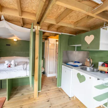 Cozy cabin interior with bed and compact kitchen, wooden ceiling and green accents.