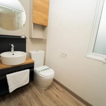 Clean bathroom with a round mirror, white sink, and toilet next to a window.