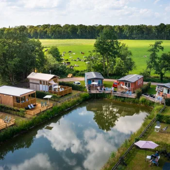 Cabins by a pond, surrounded by green fields and trees under a partly cloudy sky.