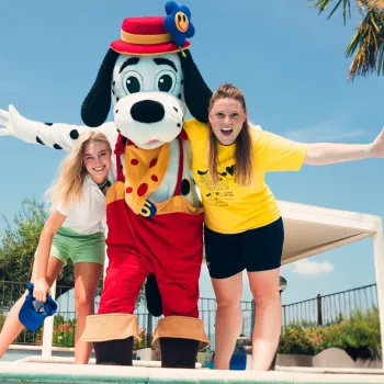 Cartoon dog mascot with two smiling women by a pool, sunny day.