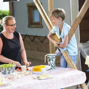 Woman and child smiling at a table with food outdoors.