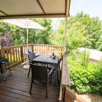 Covered patio with table and chairs, grill, umbrella, lush greenery in the background.