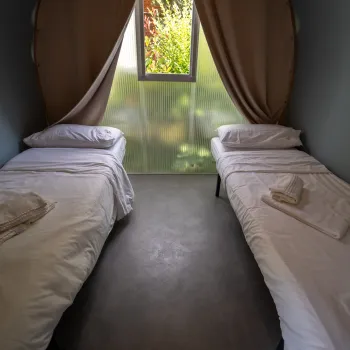 Two neatly made beds in a small room with a window and curtains.