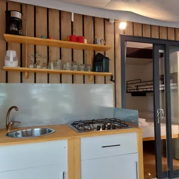 Small kitchenette with a sink, stove, and shelves, adjacent to a room with bunk beds.