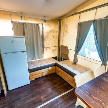 Small room with a fridge, bench seating, curtains, and wooden flooring.