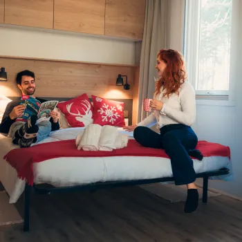 Man and woman chatting on a bed in a warmly lit room.