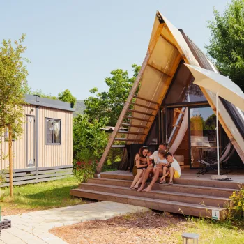 A-frame cabin with people sitting on the steps, surrounded by greenery and a small car nearby.