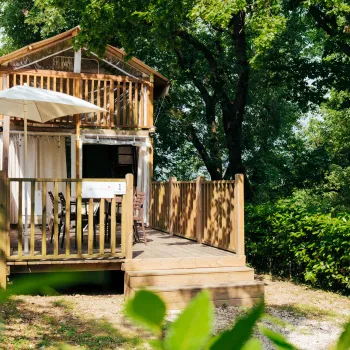 Wooden cabin with a balcony and umbrella, surrounded by lush greenery.