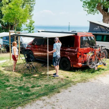 Camping scene with a red van near a lake, people setting up an awning, bikes on a rack.