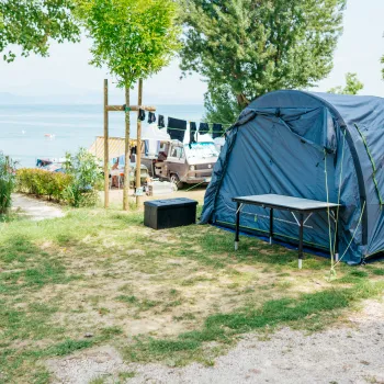 A blue tent and table on grassy campsite by a lake, trees, and parked vehicles.