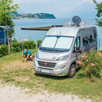 
Van aménagé garé près d'un lac avec une personne assise à proximité, surf et verdure autour.  
