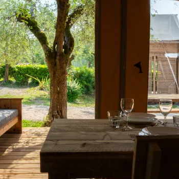 Rustic wooden table set for two, overlooking a sunny garden and wooden building.