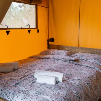 Bed inside a tent with floral bedding, two rolled towels, and soft lighting.