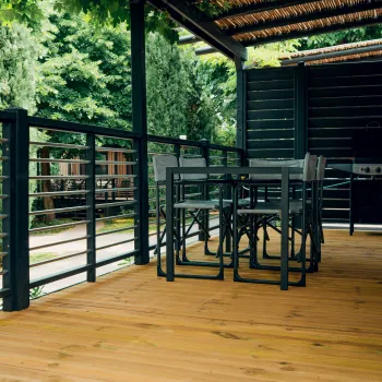 Covered wooden deck with chairs and a table, surrounded by green trees.