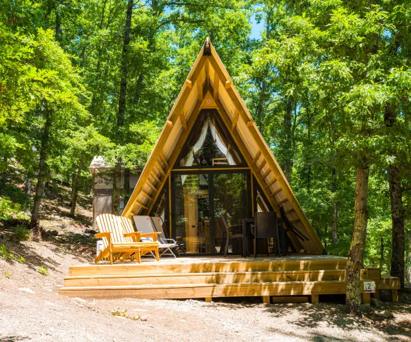A-frame cabin in a forest, with a small deck and chairs.