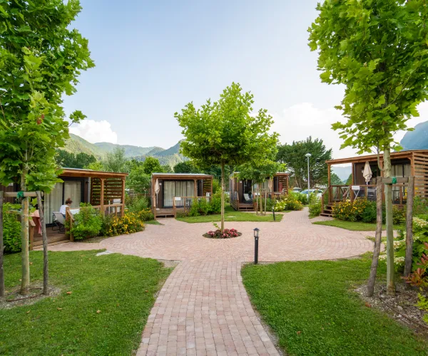 Pathway leading through a green park with cabins and trees against a mountain backdrop.