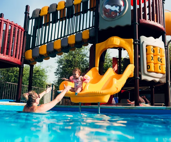 Child sliding into pool, adult reaching to catch, colorful playground backdrop.