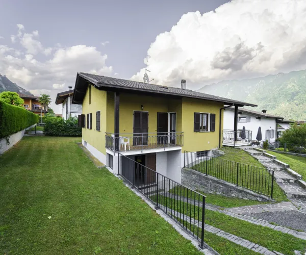Yellow house with a sloped lawn, mountain backdrop, and a cloudy sky.