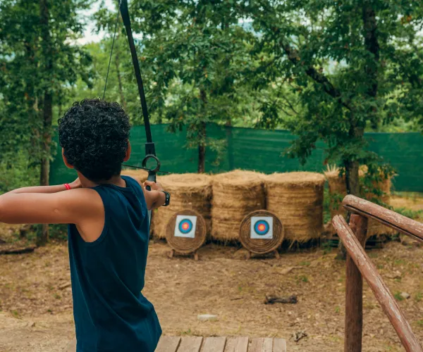 Child aiming bow at hay target in a wooded area.