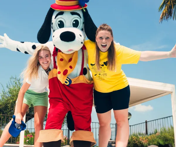 Cartoon dog mascot with two smiling women by a pool, sunny day.