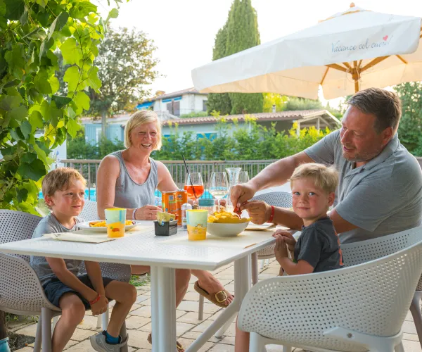 Family dining outdoors under an umbrella on a sunny day.