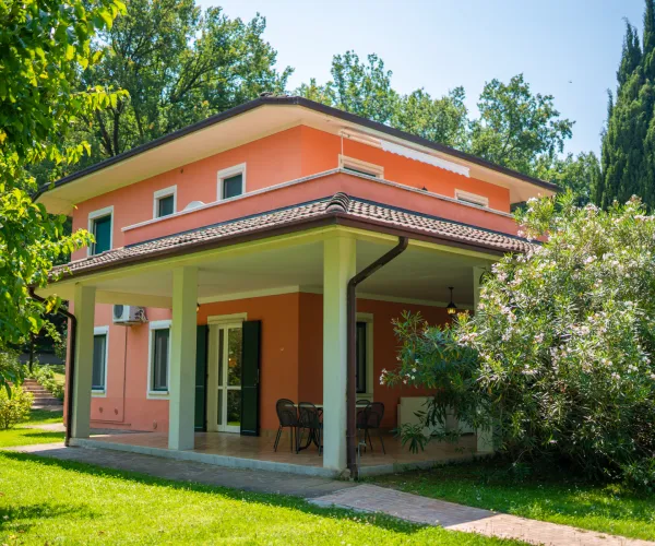 Two-storey pink house with a veranda surrounded by green trees and shrubs.
