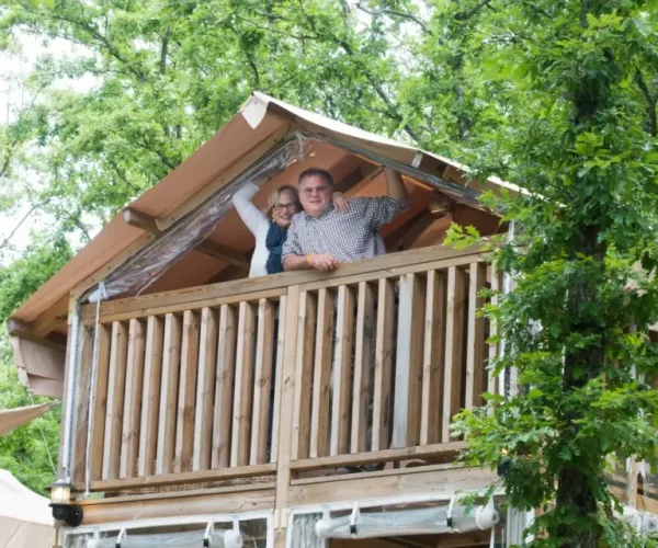 Two people waving from a wooden treehouse surrounded by green trees.