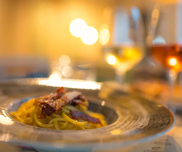 Pasta dish on a plate with two glasses of wine in a warm, softly lit setting.