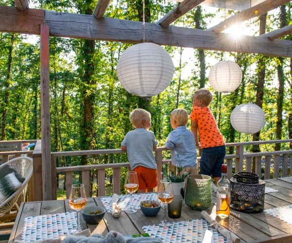 Children on a sunlit deck with lanterns, surrounded by trees.