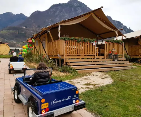 Children driving toy cars on a path near rustic cabins and mountains in the background.