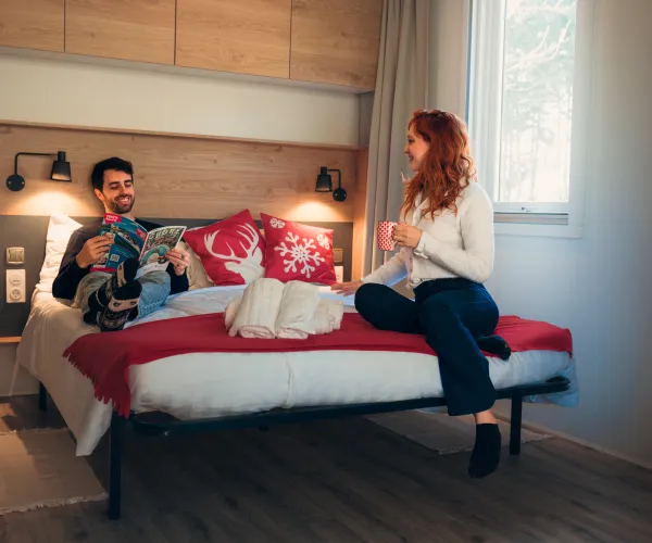 Man and woman chatting on a bed in a warmly lit room.