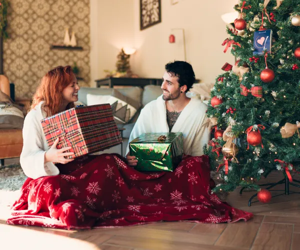 Couple in robes exchanging gifts by a Christmas tree.