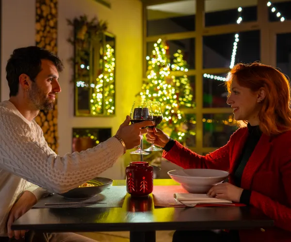 Couple toasting with wine at a cozy holiday dinner, lit Christmas tree in background.