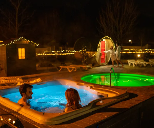 Couple relaxing in an outdoor hot tub at night, surrounded by festive lights.