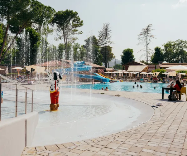 Poolside scene with fountains, people relaxing, and a water slide under a sunny sky.