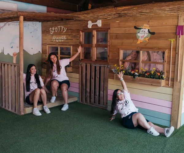 Three women smiling in front of a wooden playhouse with colorful decorations.