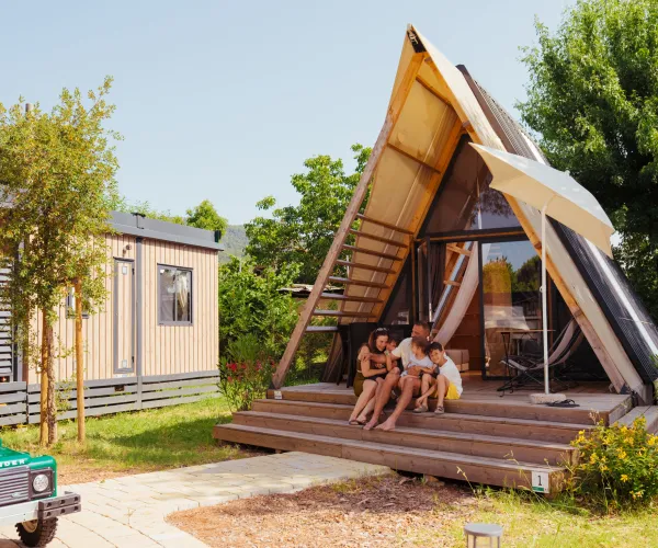 A-frame cabin with people sitting on the steps, surrounded by greenery and a small car nearby.